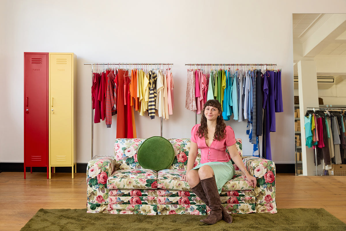 The Skinny lockers in Poppy red and Butter yellow beside rainbow-sorted garment racks in the Olga Joan fashion studio, founder seated on a floral sofa