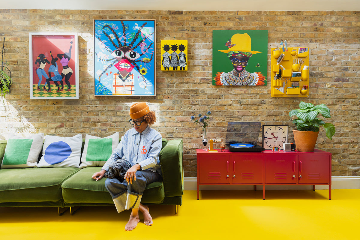 Natasha Landers on a green velvet sofa under a brick wall gallery of colourful art, beside Poppy red lockers on a yellow floor