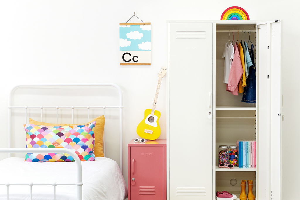 A White Twinny sits next to a Berry Shorty and a white cast iron bed in a colourful kids room. The Twinny has one door open, revealing a bright collection of kids clothes and toys, and there is a rainbow pillow on the bed.