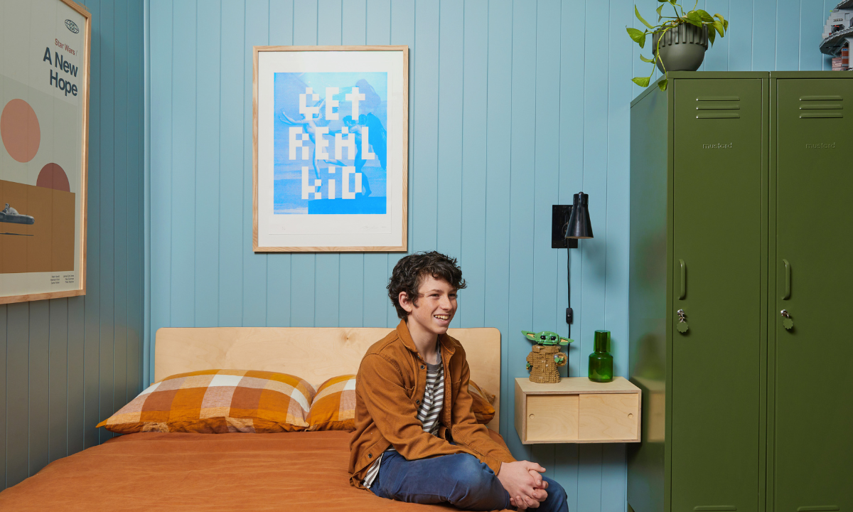 Dylan smiles as he sits on his bed. There is an Olive Twinny behind him and rust-coloured linens on the bed.