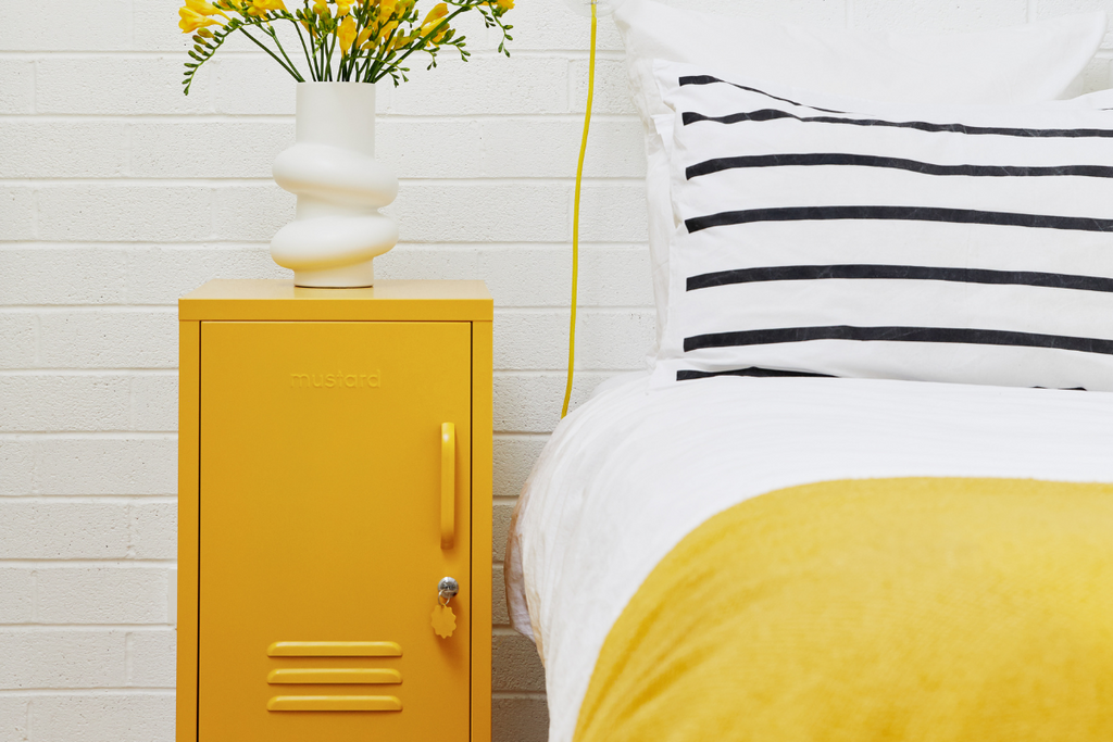 A Mustard yellow Shorty locker sits next to a bed dressed in Mustard linens with a black and white striped pillow. There is a vase of yellow flowers on top of the locker.