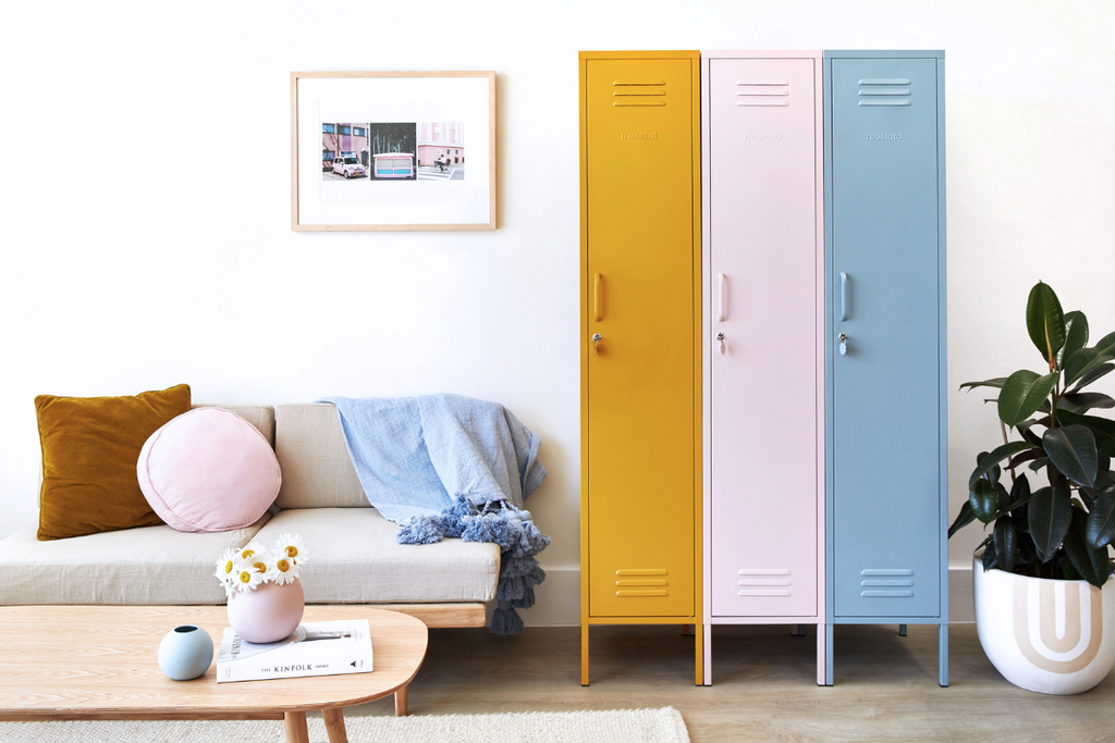 Three Mustard Made Skinny lockers stand side by side. From the left, the first is Mustard, then Blush then Ocean. To the left is a sofa with a wooden, timber frame, beige padding and cushions of various shapes and colours sit on top. 