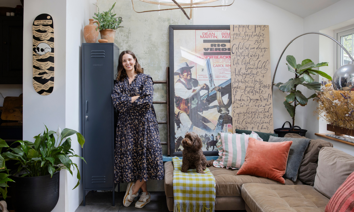 Clare Watson wears a flowing floral dress and sneakers as she leans against a Slate Skinny Mustard Made locker in her living room. She is surrounded by plants, artworks and throw cushions and there is a small dog on her couch.