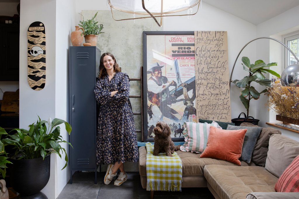 Clare Watson wears a flowing floral dress and sneakers as she leans against a Slate Skinny Mustard Made locker in her living room. She is surrounded by plants, artworks and throw cushions and there is a small dog on her couch.